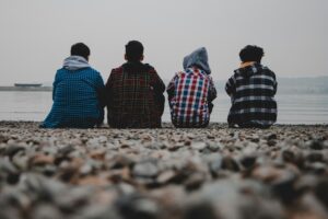 Four teenage boys sit together on a pebble beach looking out to sea, their backs to the camera